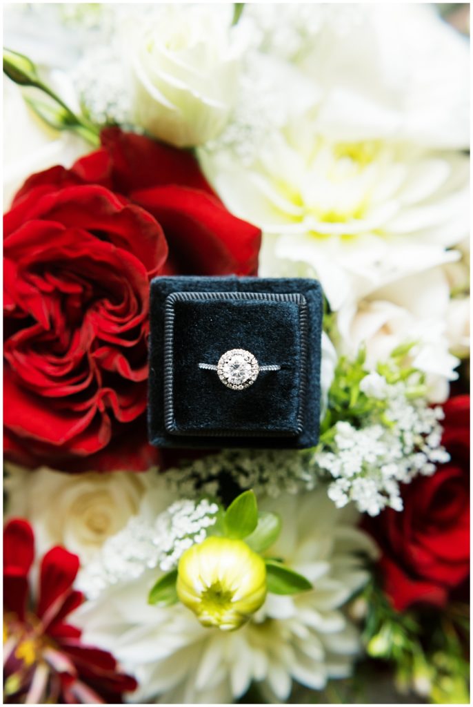 Engagement ring in a black velvet box surrounded by red and white flowers.