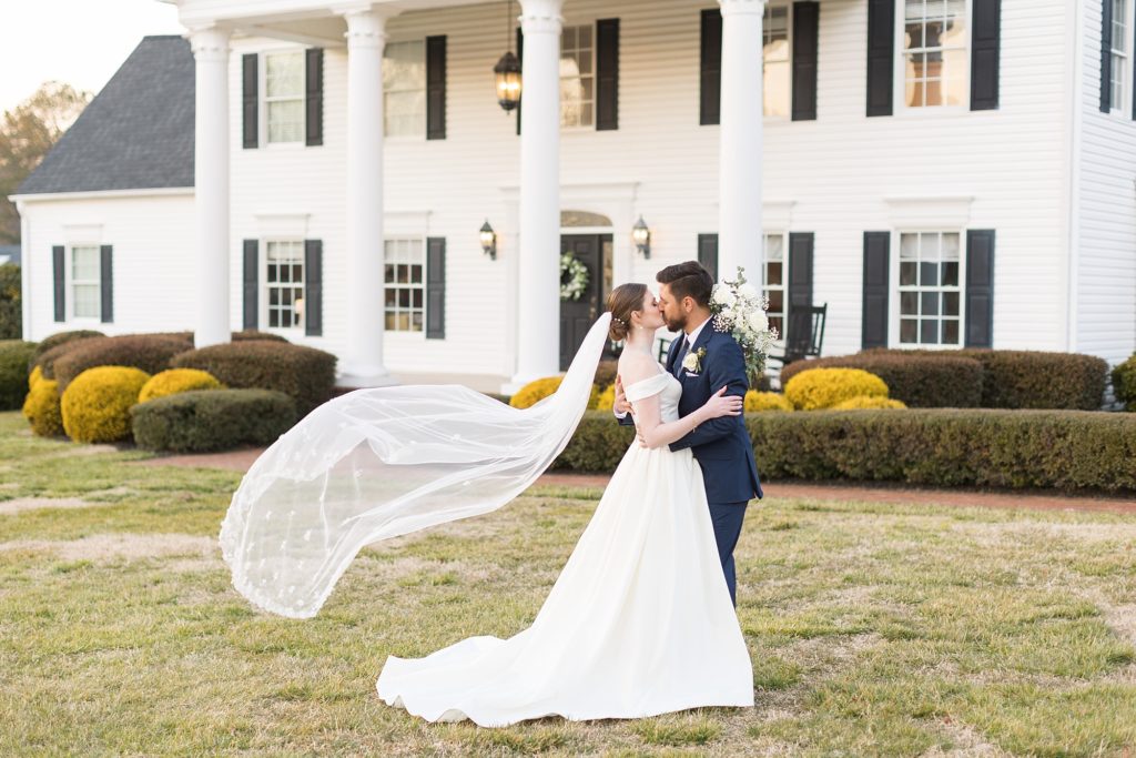 The bride and groom share a kiss in front of a classic white house as her veil flies in the wind | Raleigh Wedding Photographer