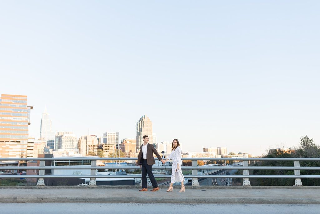 Couple walking over bridge with city skyline | Downtown Raleigh Engagement Photoshoot | Raleigh NC Wedding & Engagement Photographer | Sarah Hinckley Photography 