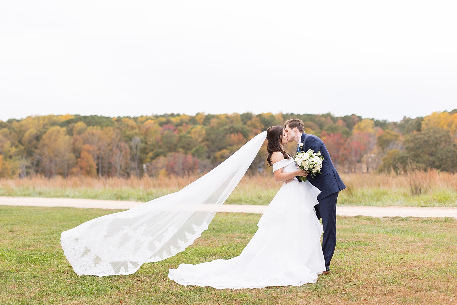 Bride and groom kissing with veil blowing in the wind | Fall Wedding at The Meadows in Raleigh | Raleigh NC Wedding Photographer