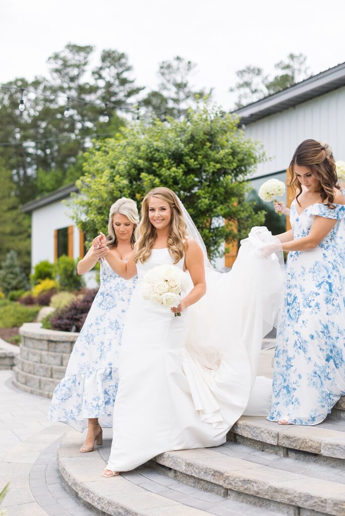 Bride walking down stairs outside venue holding white flower bouquet and bridesmaid holding peonies | Classic blue and white wedding | The Evermore Wedding | The Evermore Wedding Photographer | Raleigh NC Wedding Photographer
