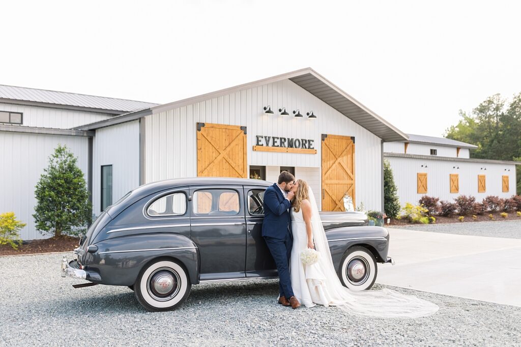 Bride and groom kissing by classic automobile in front of venue | The Evermore Wedding | The Evermore Wedding Photographer | Raleigh NC Wedding Photographer