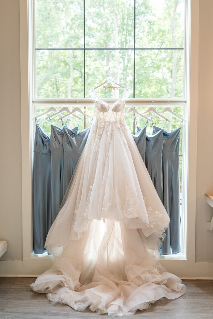 Bridal dress hanging in front of bridesmaids dresses in front of window | Blue and white Wedding | Carolina Groves Wedding | Carolina Groves Wedding Photographer | Raleigh NC Wedding Photographer